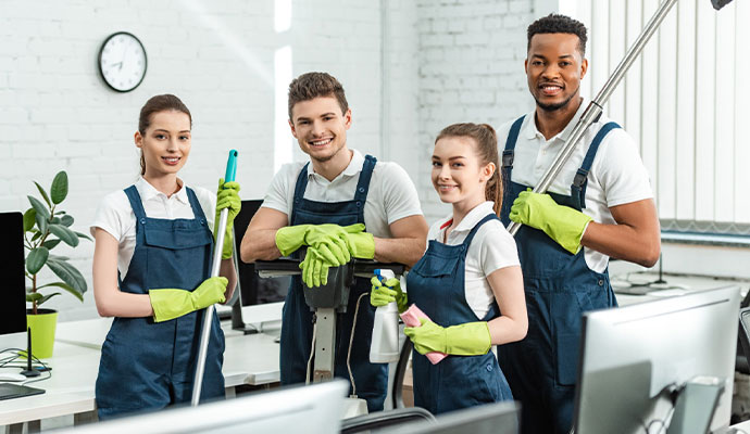 A professional team of four cleaners in matching blue uniforms and green gloves holding cleaning equipment in a bright, clean office.