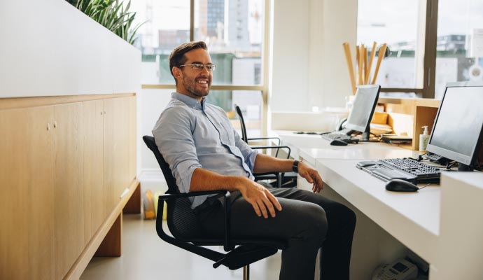 Office employee seated at ergonomic desk with dual monitors, keyboard, and organized supplies in front of large windows in an imrpoved air environment