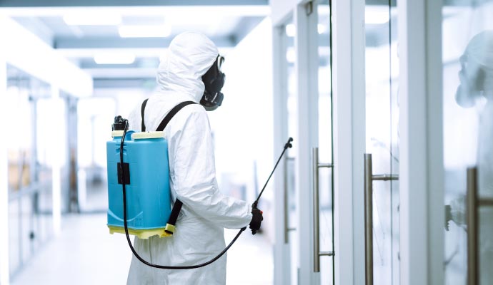 Technician in hazmat suit and gas mask disinfecting a glass door using a blue sprayer unit inside a sanitized facility