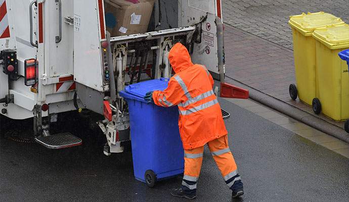 A sanitation worker in a bright orange safety uniform loading a blue waste bin into a modern white garbage truck.