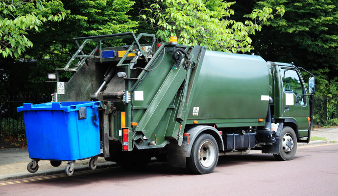 A green garbage truck lifting a large blue waste bin on a city street near lush trees