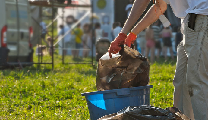A cleanup worker in orange gloves removing a full trash bag from a blue bin during a crowded outdoor festival