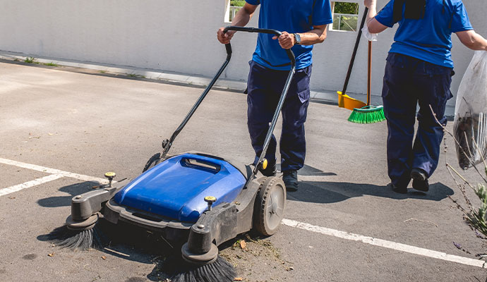 A cleaning professional in a blue uniform using a industrial walk-behind floor sweeper on an outdoor asphalt parking area.