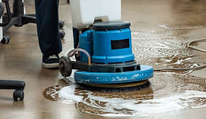 A blue industrial floor scrubbing machine being used with water and cleaning solution to restore a concrete office floor.