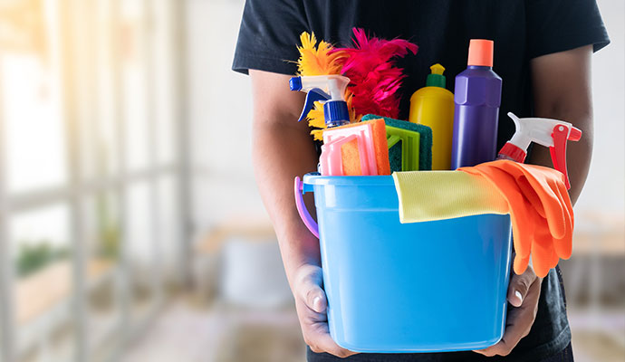 A professional holding a blue cleaning caddy filled with various janitorial supplies, including spray bottles, sponges, brushes, and protective gloves.