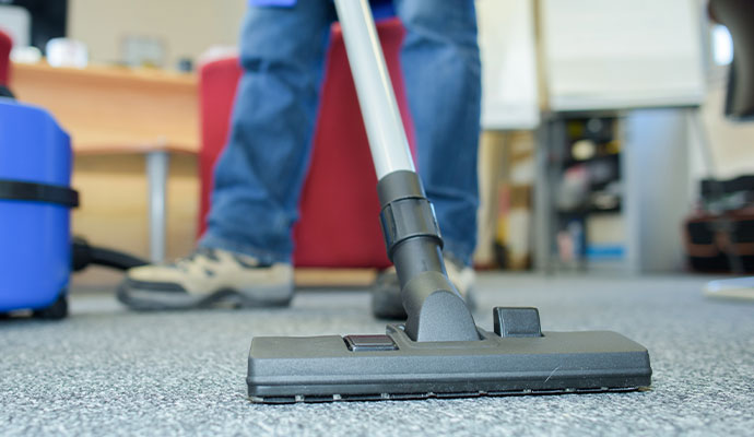 A close-up of a professional cleaner using a heavy-duty vacuum attachment to deep clean grey commercial carpeting in an office setting.