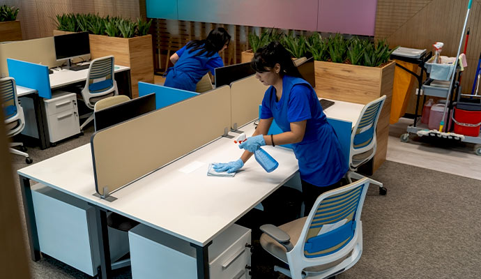 Two cleaners in blue uniforms wiping down office desks.