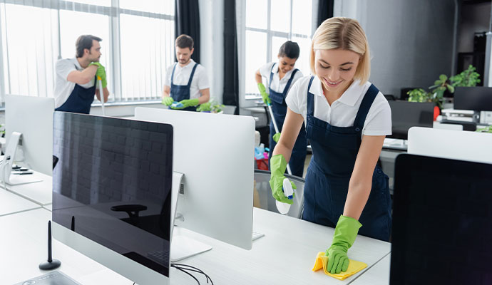 A team of professional cleaners in blue overalls and green gloves sanitizing white workstations and mopping floors in a bright, modern office in DeSoto.