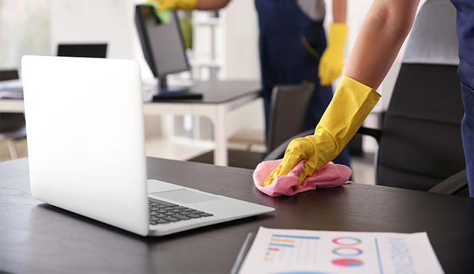 A close-up of a cleaner in yellow gloves wiping down a dark office desk and workstation with a pink cloth to remove accumulated dust.