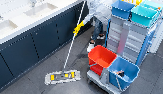 A professional cleaner in a white coat and yellow gloves mopping a dark grey tiled restroom floor next to a fully stocked janitorial cart with colorful buckets.