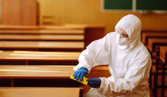 A worker in a hazmat suit sanitizing desks in a classroom.