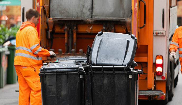 Workers loading waste bins into an orange garbage truck.
