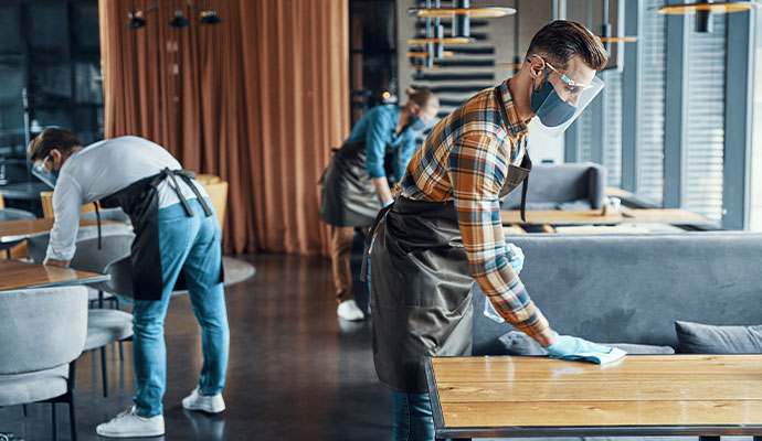 Three professional cleaners wearing face shields and masks sanitizing tables and seating in a modern DeSoto restaurant.